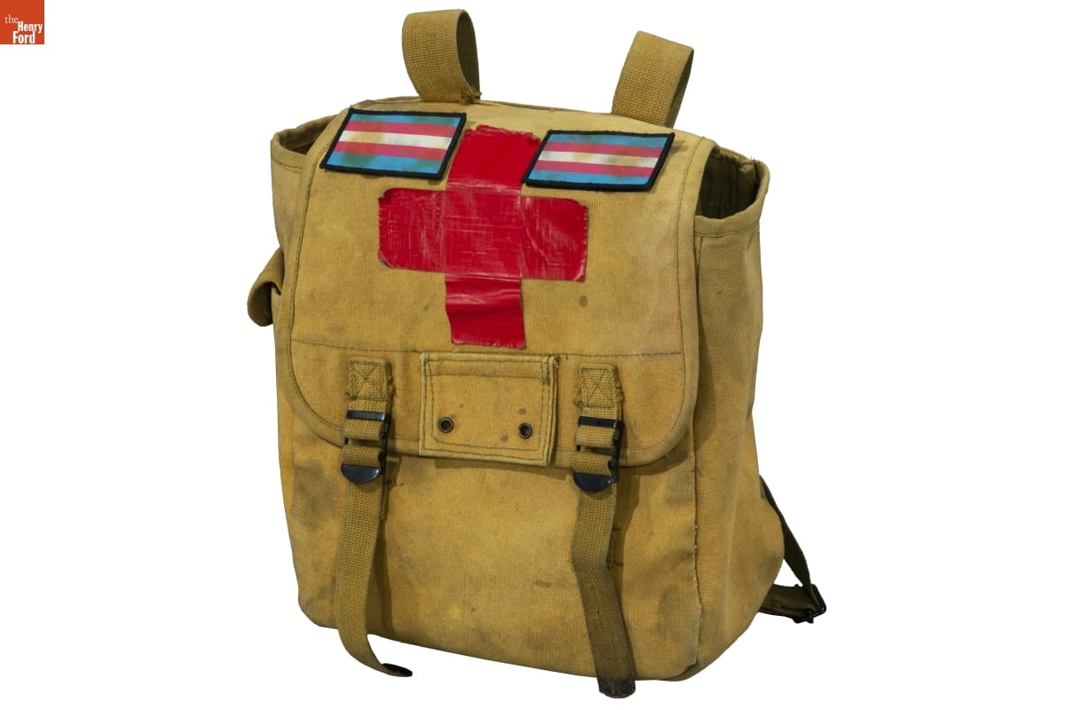 Khaki-colored knapsack or backpack with cross made out of red duct tape and striped patches at top