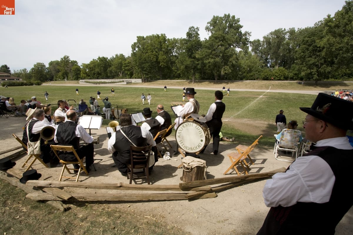 A group of people with musical instruments sit and stand in a circle next to a baseball field and spectators