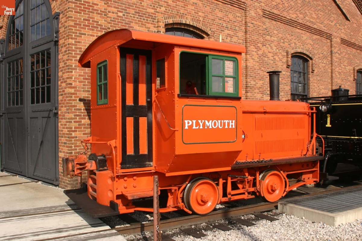 Small orange locomotive on train tracks in front of brick building